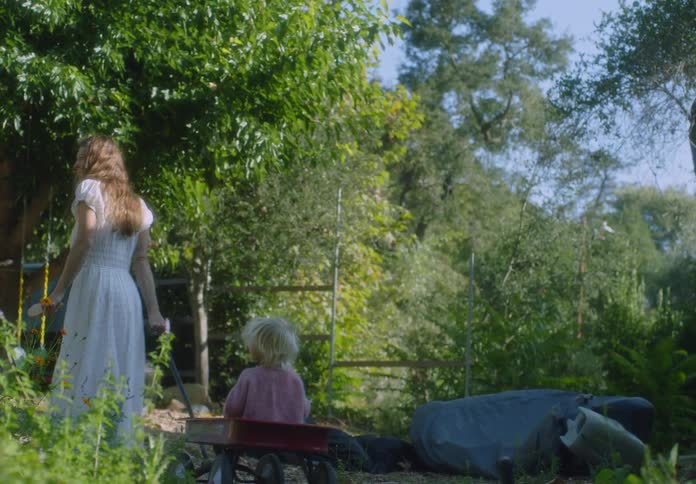Woman wearing a long white dress pulls her little girl in a wagon on a rural forested mountain