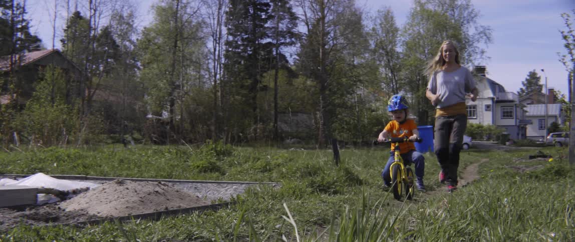 Mother playfully chasing her young son while he practices riding a bike in nature.