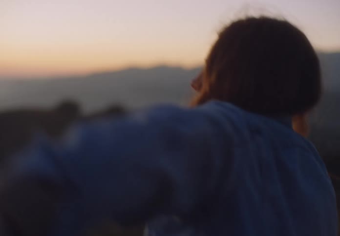A young woman with long hair dances freely on a grassy hilltop during a golden sunset wearing a denim jacket and white top as the horizon glows with orange and purple light over distant rolling mountains