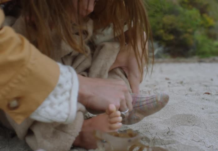 Mother helps daughter remove her shoes and socks while the girl sits on the sandy beach on an autumn day