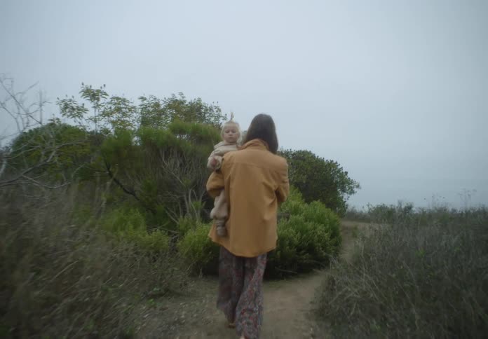 A woman wearing a yellow jacket and floral dress walks along a dirt path on a foggy cliff while carrying a young child in a tan fleece outfit who holds a piece of green foliage