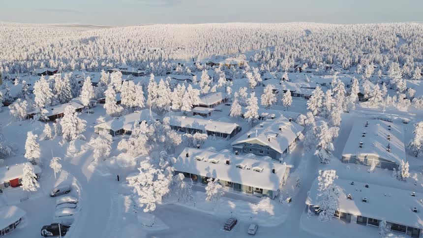 This aerial footage showcases a cluster of snowcovered buildings nestled within a vast frozen forest landscape the camera slowly pans across the quiet winter settlement revealing rooftops and trees blanketed in deep white snow under a clear bright sk