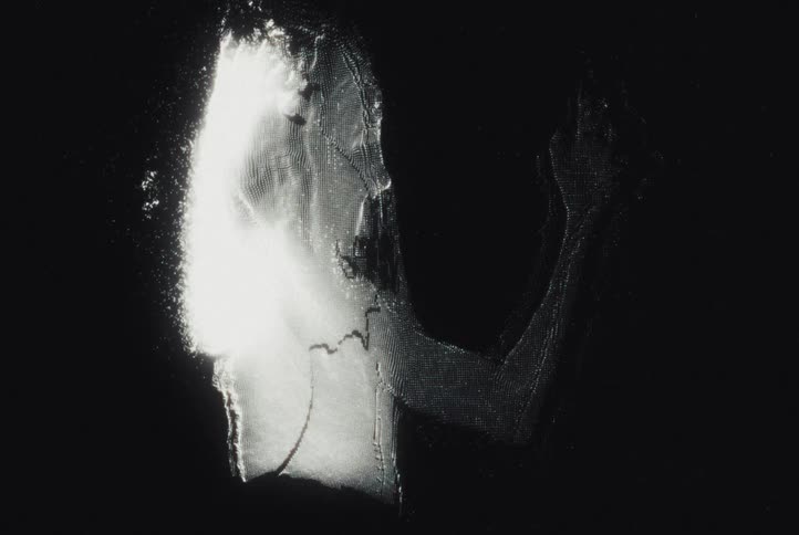 A glowing abstract silhouette of a person raises a hand against a dark background their form dissolving into shimmering particles of light