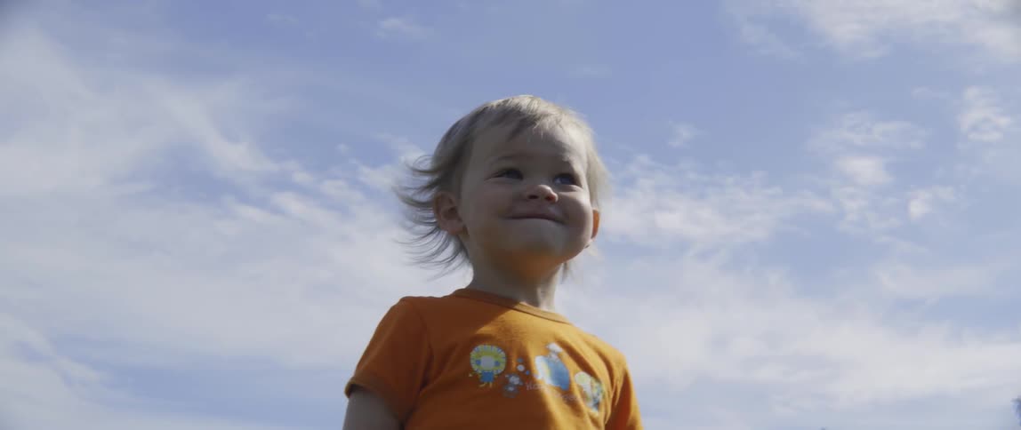 A toddler boy is standing outside on a sunny day and smiling
