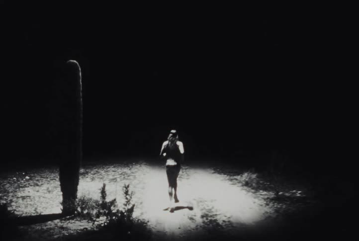 Man with long hair passes a cactus while running on a dirt road in the desert at night