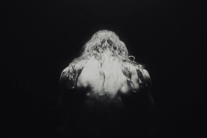 Shirtless person with head tilted downward against a dark background