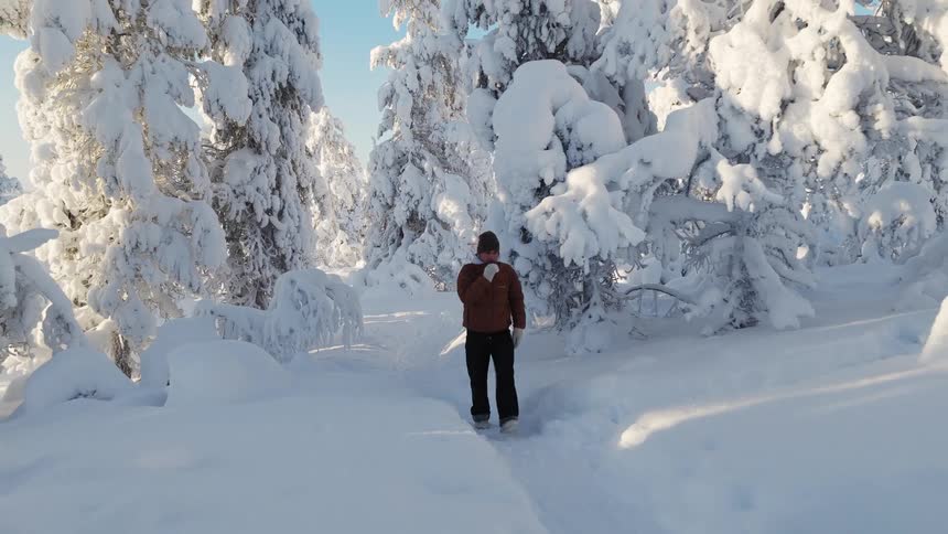 A man in a brown jacket and knit cap walks along a narrow path through a dense forest of trees heavily weighed down by thick white snow
