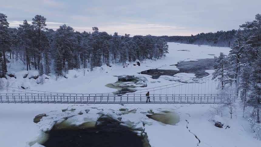 River in the forest flows through broken ice as a hiker walks across a footbridge near a rural house
