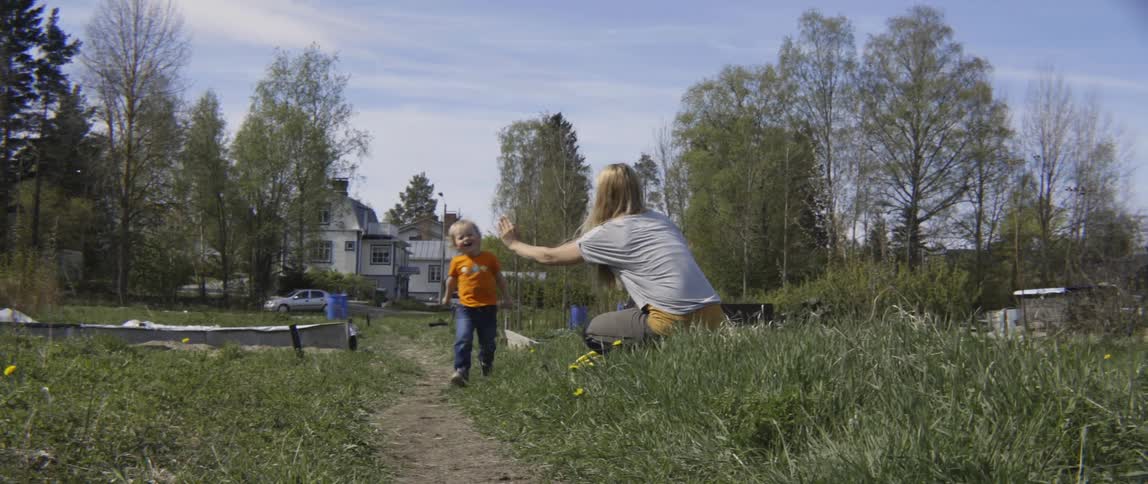 A young toddler boy runs through a grassy field toward a woman standing outdoors.