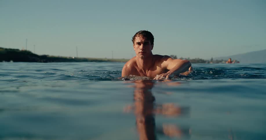 A man swims on a surfboard in the ocean near other surfers with his head above the water while looking toward the horizon