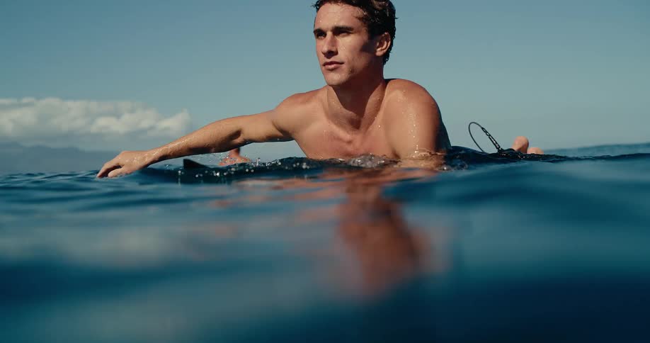 A man swims in the ocean on his surfboard with his head above the water near other surfers while looking toward the horizon
