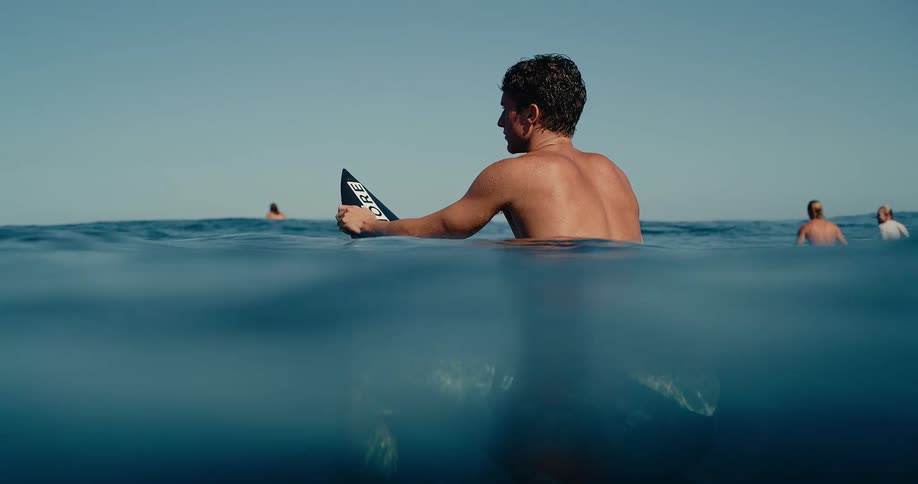 A surfer sits on his board in the ocean glancing over his shoulder looking at other surfers while waiting for waves under a clear blue sky