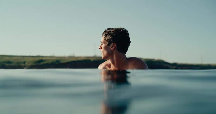 A man swims in the ocean on his surfboard with his head above the water while looking toward the horizon