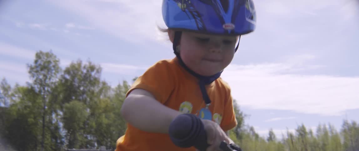 A young toddler boy walks his small bicycle backwards through the grass in nature.