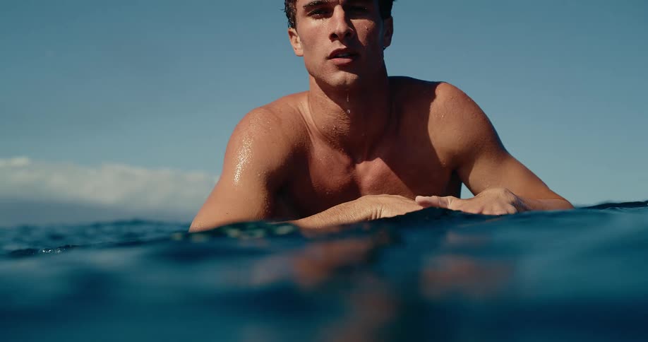 A shirtless male surfer floats on his board in the ocean water