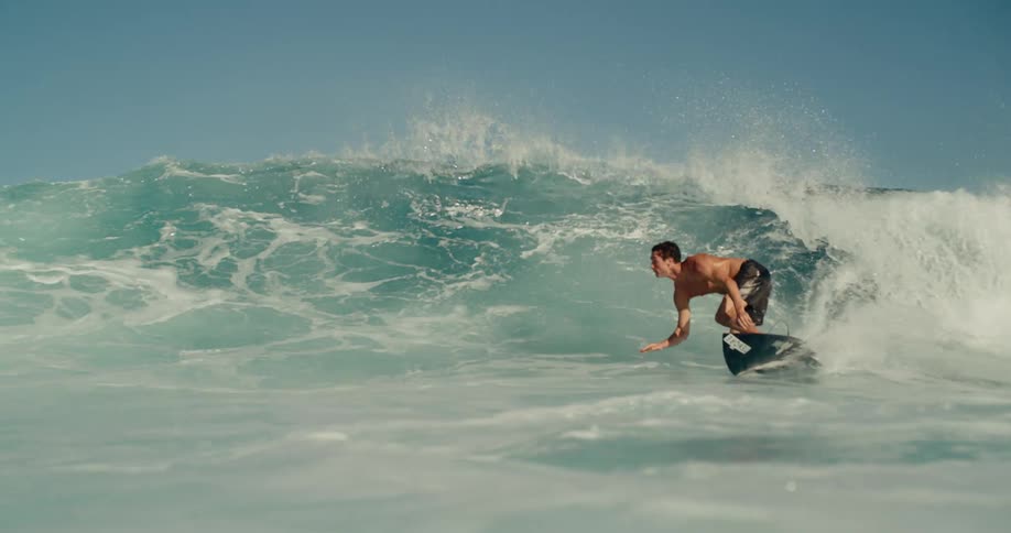 A man wearing black shorts rides a large ocean wave on a surfboard under a clear blue sky before being covered by the white spray of the water