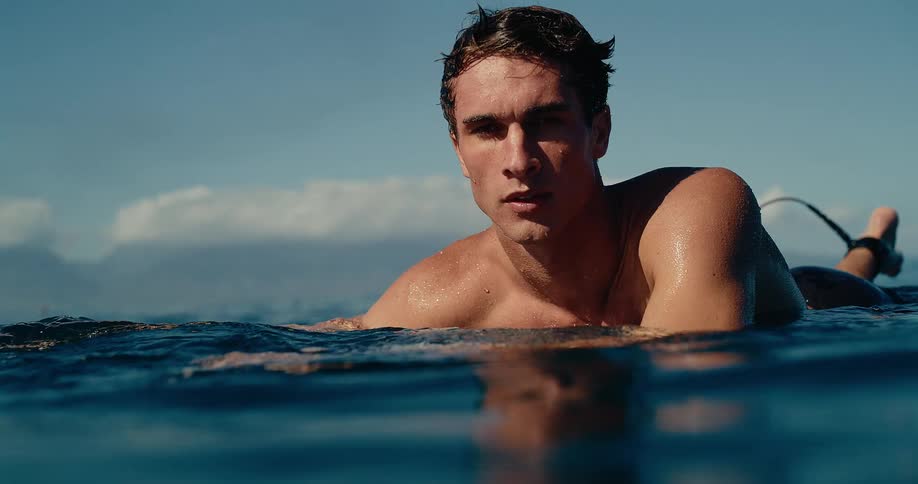 A shirtless male surfer lays on his board waiting for a wave