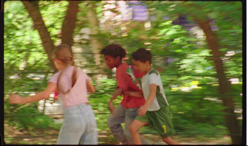Children run through a park while kicking a soccer ball surrounded by trees and greenery with sunlight filtering through the leaves