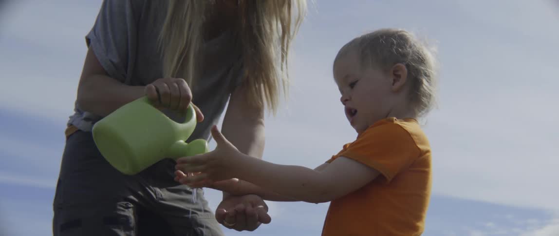 Woman pouring water from a pitcher onto a toddler's hands outdoors