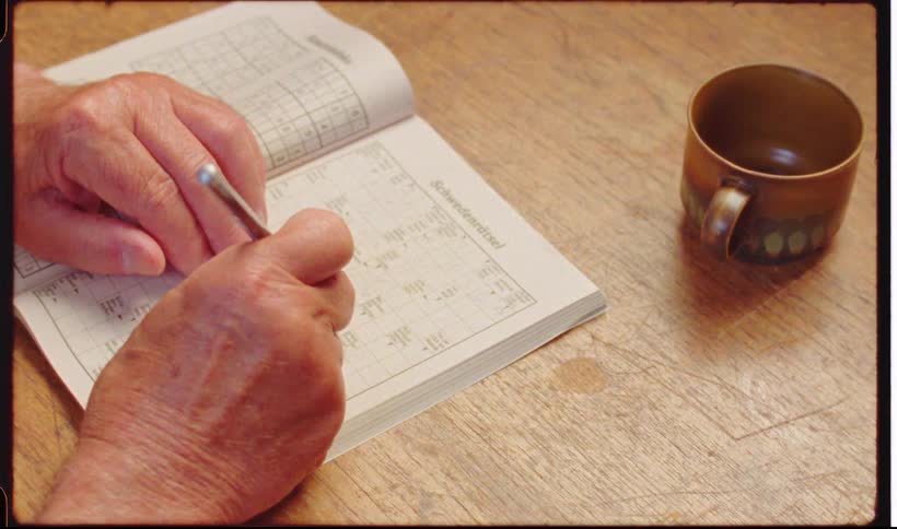 Man sitting at a wooden table works on a crossword puzzle