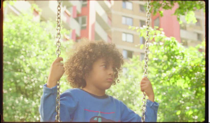 A young boy with curly hair sits quietly on a metal swing set in a sunny park with leafy green trees and a tall residential building in the background
