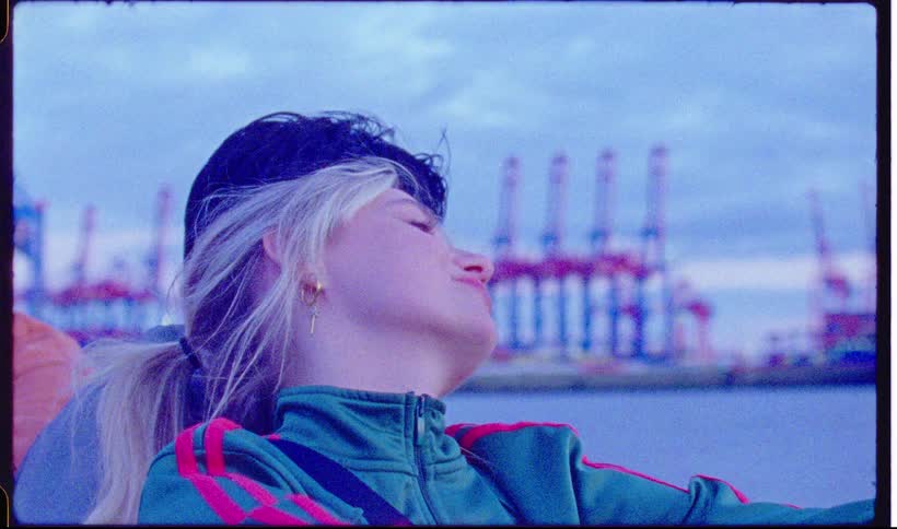 A woman wearing a jacket and beret leans back against another person smiling near a harbor with cargo cranes and water in the background