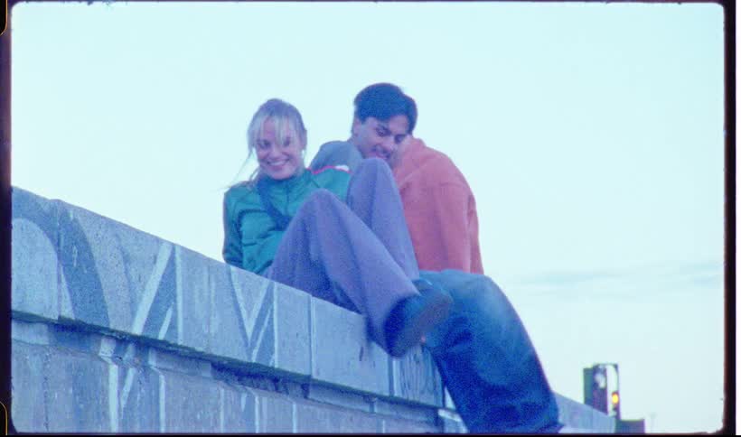 A group of friends in casual clothes run toward a stone wall and climb up to sit together while talking and smiling against a pale sky with a red boat and harbor visible in the distance