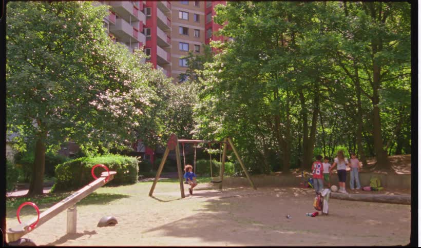 Group of children playing together in a sunny neighborhood park playground surrounded by trees and apartments