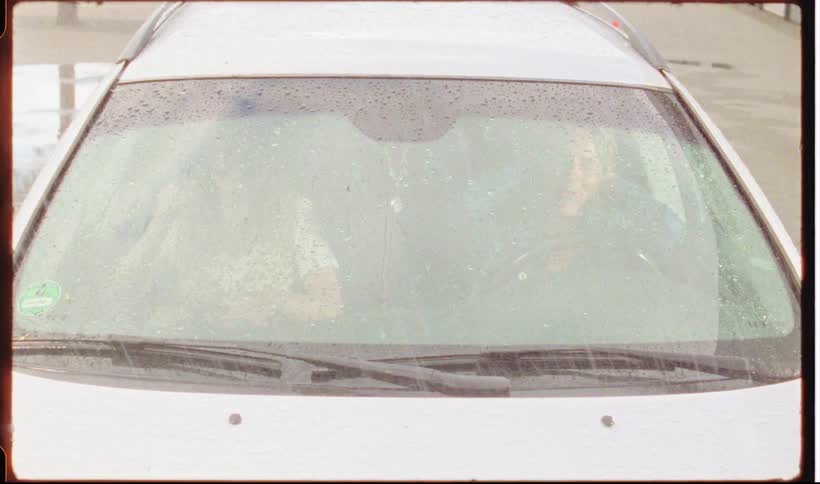 A view through a wet car windshield shows two people sitting inside a white vehicle during a rain shower