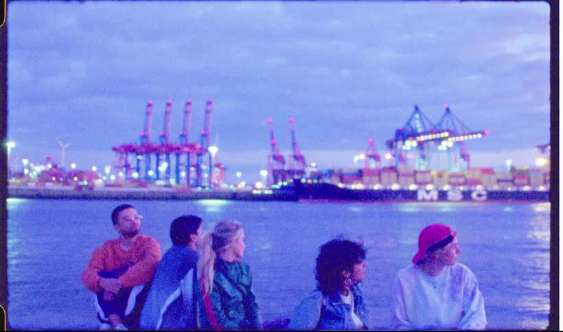 Five people sit by the water looking toward a harbor with large cargo cranes and ships in the background at dusk