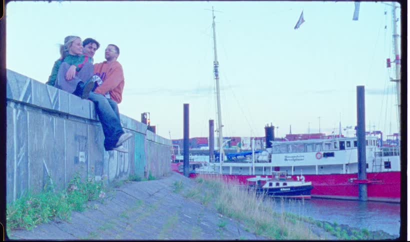 A group of friends run toward a concrete wall and sit on top of it next to a harbor with large red ships and green water under a clear sky
