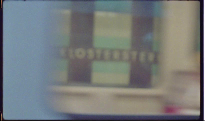 A view through a vintage camera lens shows a train with a blue and white exterior as it passes by a station platform with blurry figures in the background