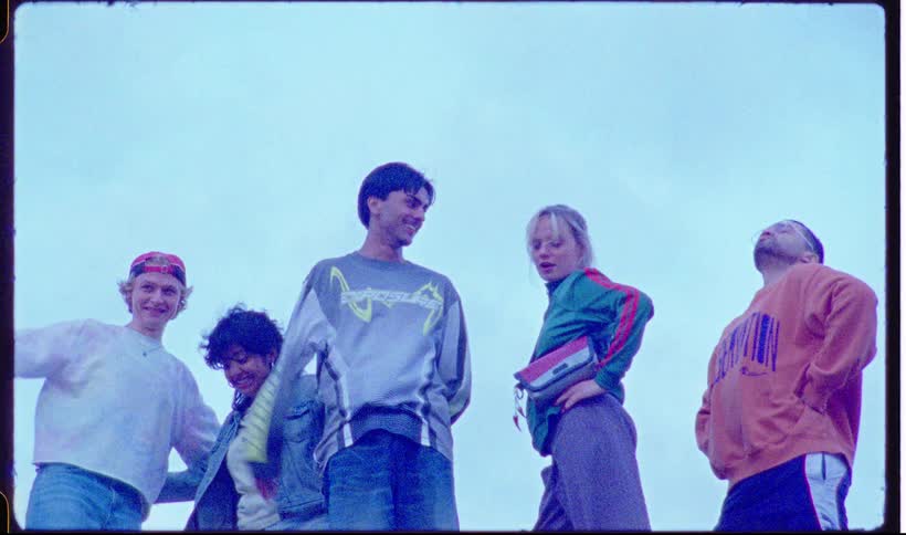 Five young adults stand outdoors against a bright sky posing and moving with a playful retro energythe group transitions from upbeat dancing and saluting to a relaxed smiling stance as they look down toward the camera