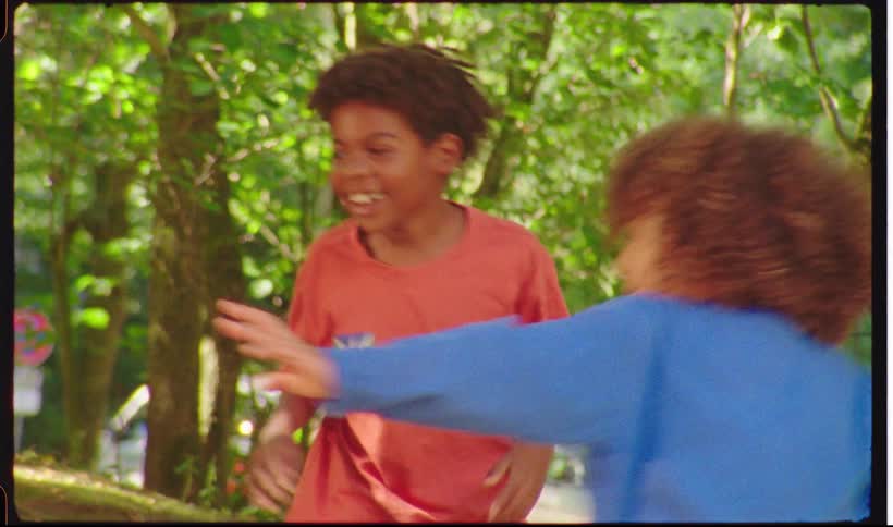 A group of young children are seen laughing and playing together outdoors in a sunlit wooded area the vintage film style captures their joyful expressions as they run around and interact near a fence