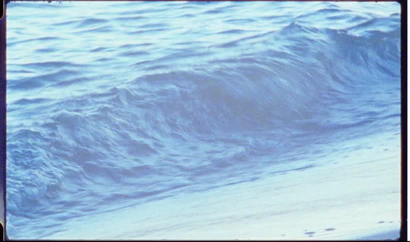 A view through a vintage camera lens shows gentle blue waves with white foam rolling onto a sandy beach during a bright day