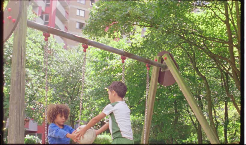 Two boys stand near a swing set in a sunny park with one holding a white soccer ball after a brief exchange the boy in the blue shirt hands the ball to the other child who then runs out of the frame