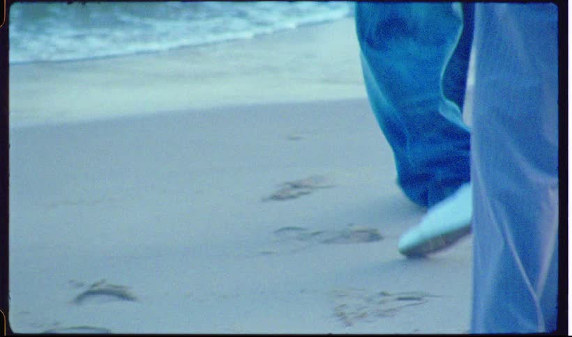 A lowangle shot of people in 1990sstyle athletic wear walking and dancing along a sandy shoreline the footage has a grainy vintage film aesthetic with bluetoned waves crashing in the background as the group moves across the wet sand