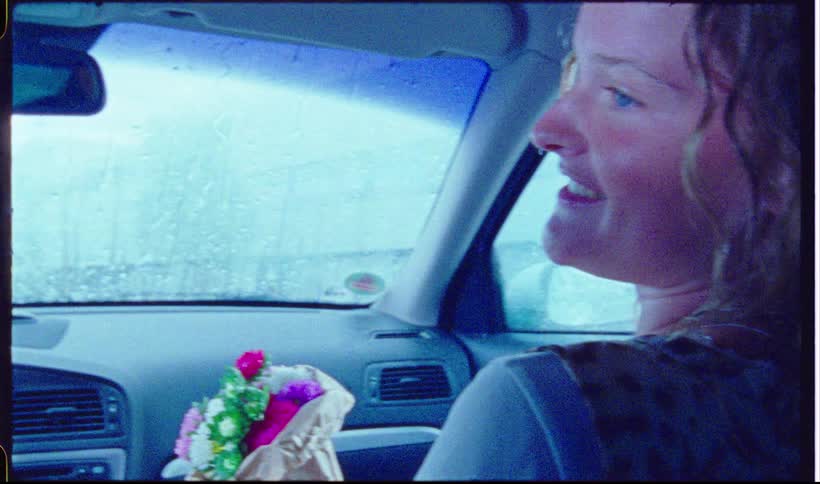 A woman with wavy hair sits in the drivers seat of a car on a rainy day smiling as she looks toward the passenger side a colorful bouquet of flowers rests on the dashboard while raindrops cover the windshield
