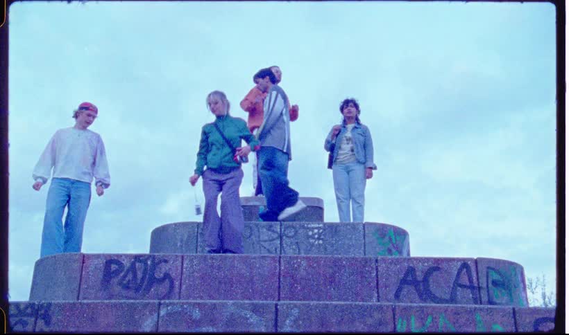A group of young people hang out and move around on top of a tiered graffiticovered stone structure the footage has a vintage grainy film aesthetic with the group posing and taking photos against a cloudy sky
