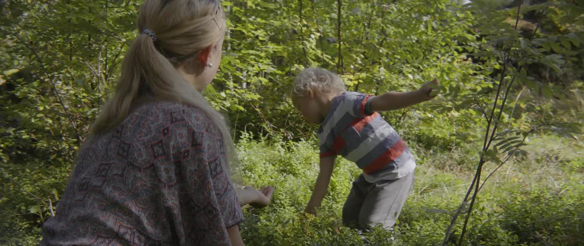 A young toddler boy hands green leaves to his mother while sitting in grass.