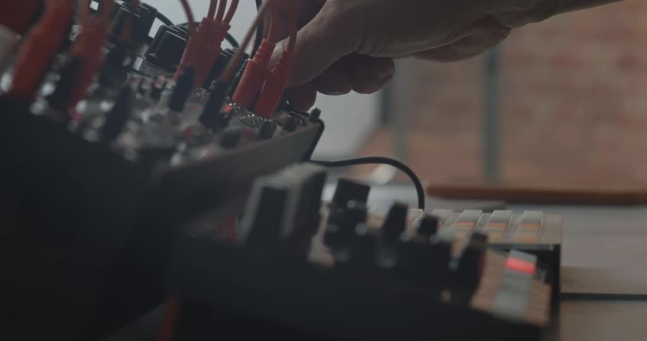 A persons hand adjusts buttons on a modular synthesizer on a wooden table while a grid controller sits blurred in the foreground of a dimly lit studio