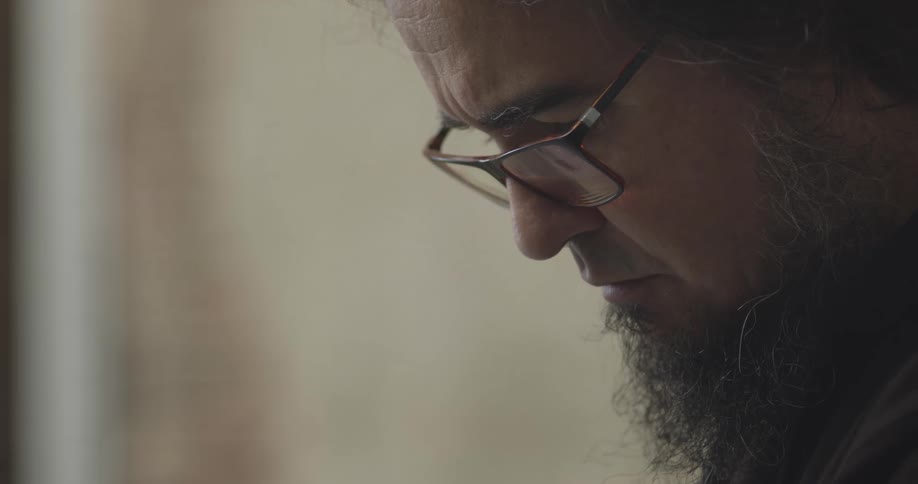A man in profile close up with glasses and a long beard looks downward in concentration indoors