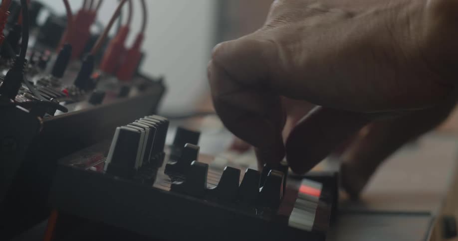 A man uses his fingers to press keys and adjust various knobs on a portable electronic synthesizer while other hardware units and orange patch cables sit connected in a shallow depth of field studio setting