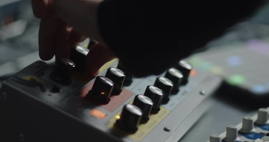 A close view from the side shows a person using fingers to adjust various black knobs on a silver electronic device with orange lights in a dark room