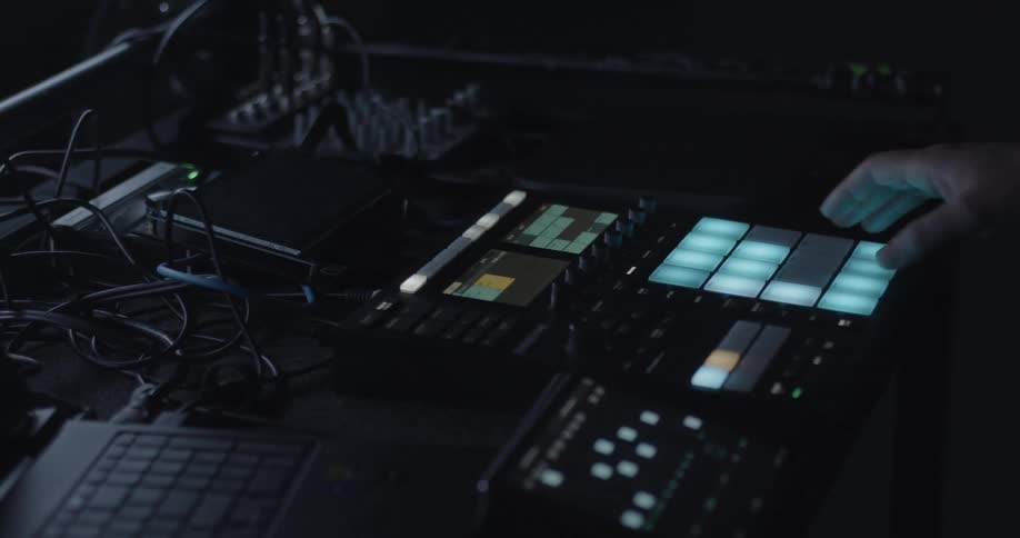 Music producer creating electronic music using a pad controller and equipment on a desk in a dark studio workspace