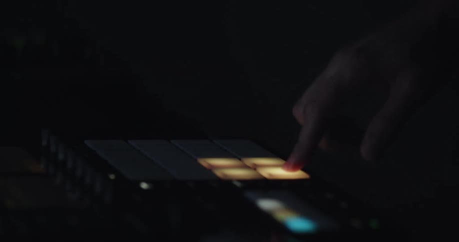 A hand presses illuminated pads on a midi controller in a dark environment the pads change colors from blue to orange as they are triggered during the musical performance