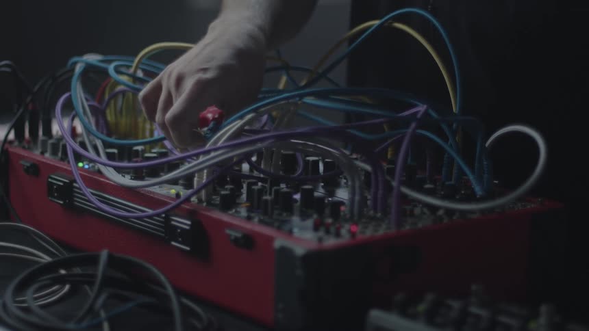 A person adjusts knobs and cables on an electronic music synthesizer with multiple colorful wires connected