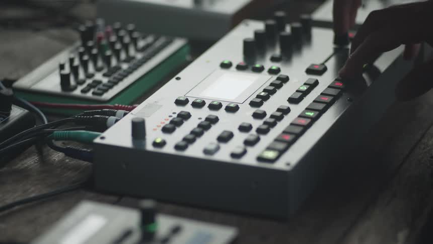 A persons hands adjusting the knobs and pressing buttons on an electronic music sequencer it captures a closeup atmospheric view of a live hardware synthesis setup on a wooden surface
