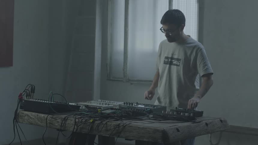 A man adjusts settings on electronic synthesizers and audio equipment placed on a wooden table