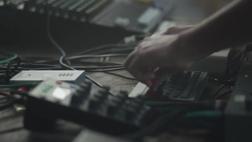 A persons hands are shown adjusting knobs and controls on a variety of electronic music equipment and synthesizers the setting appears to be a dimly lit studio or live performance space filled with tangled patch cables and mixers